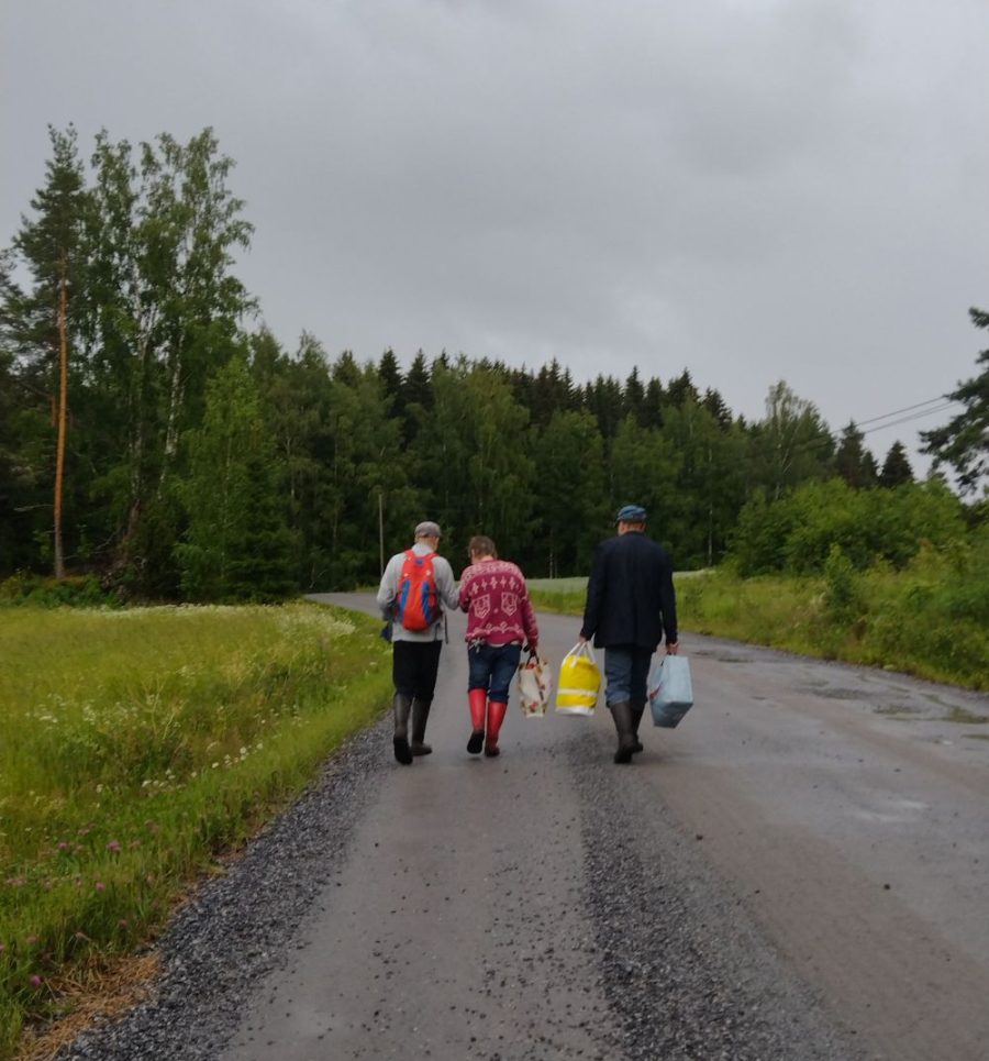 Kolme ihmistä kävelee hiekkatiella maalaismiljöössä